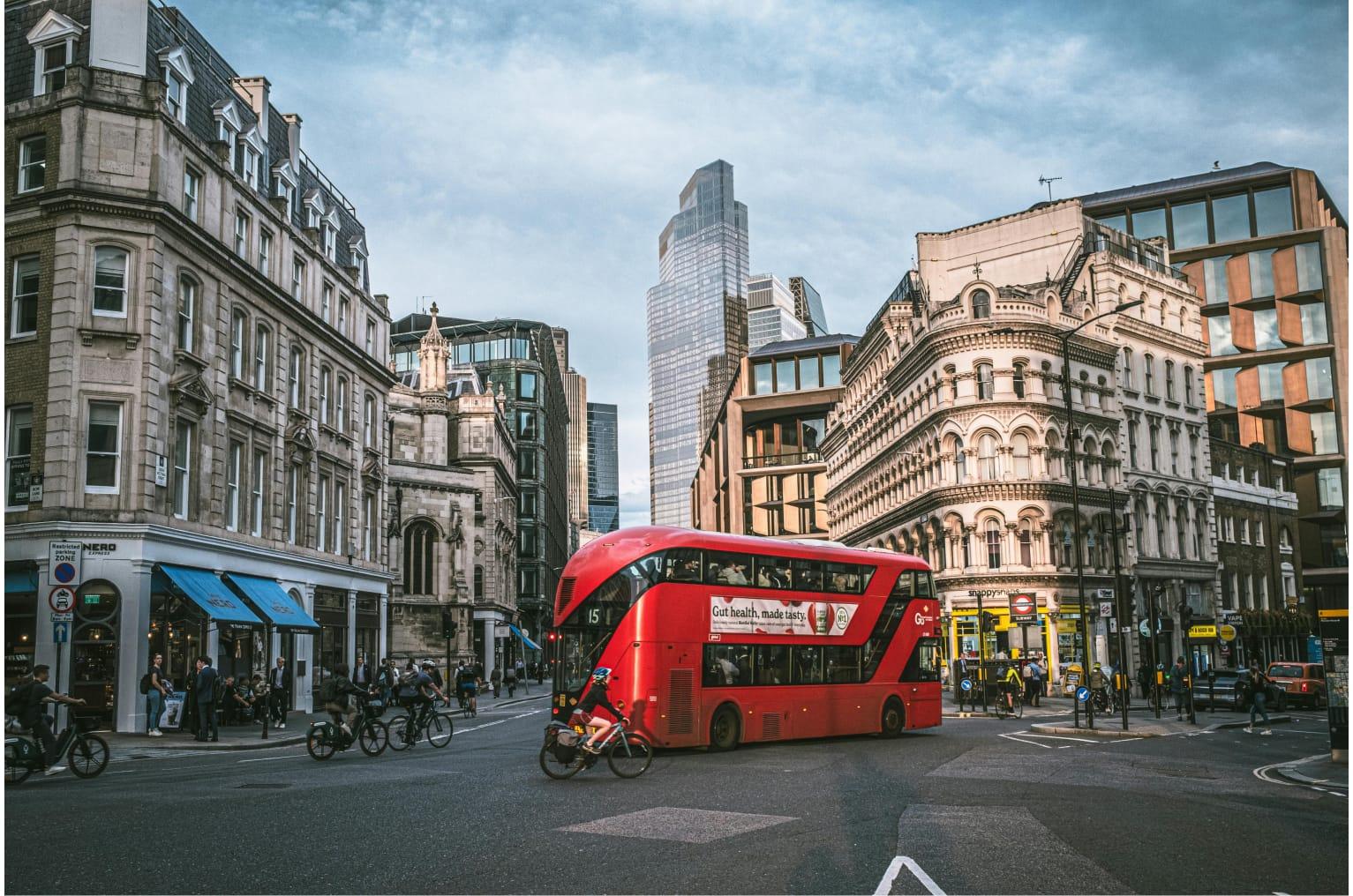 A red double deck bus is centre frame. In the background there are buildings and skyscrapers. The sun is out and the weather is nice.