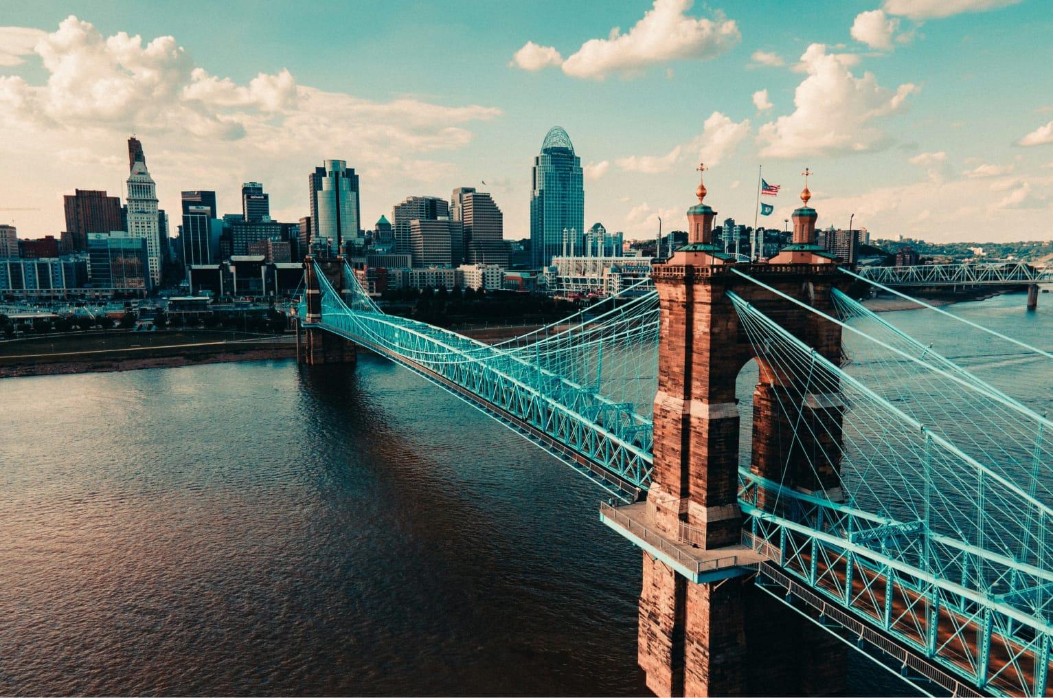 The John A. Roebling Suspension Bridge and the Ohio River with a skyline of buildings in the background - the sky has an orange tinge.