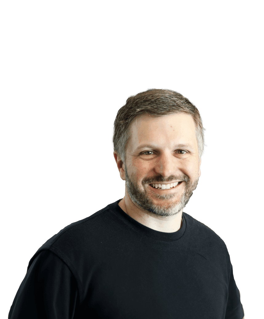 A man stands smiling into the camera, wearing a black jumper in front of a blurred background.