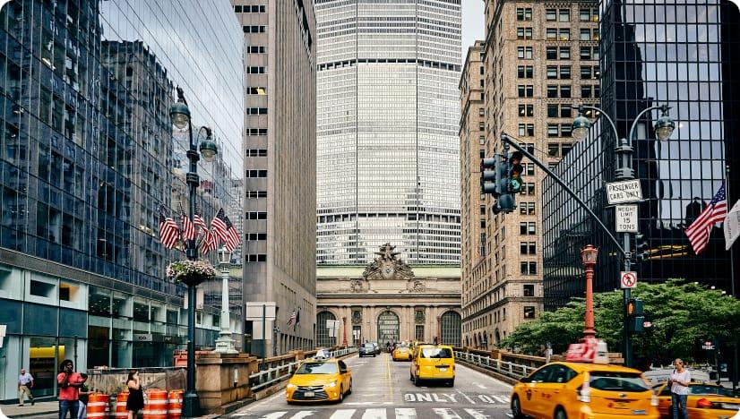 New York yellow taxis drive in between tall buildings with Grand Central Station visible in the background.