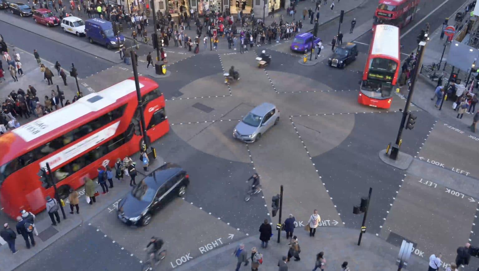 Aerial shot of a red London bus turning at a junction next to a populated street of shoppers. Black cabs wait for the bus to turn.