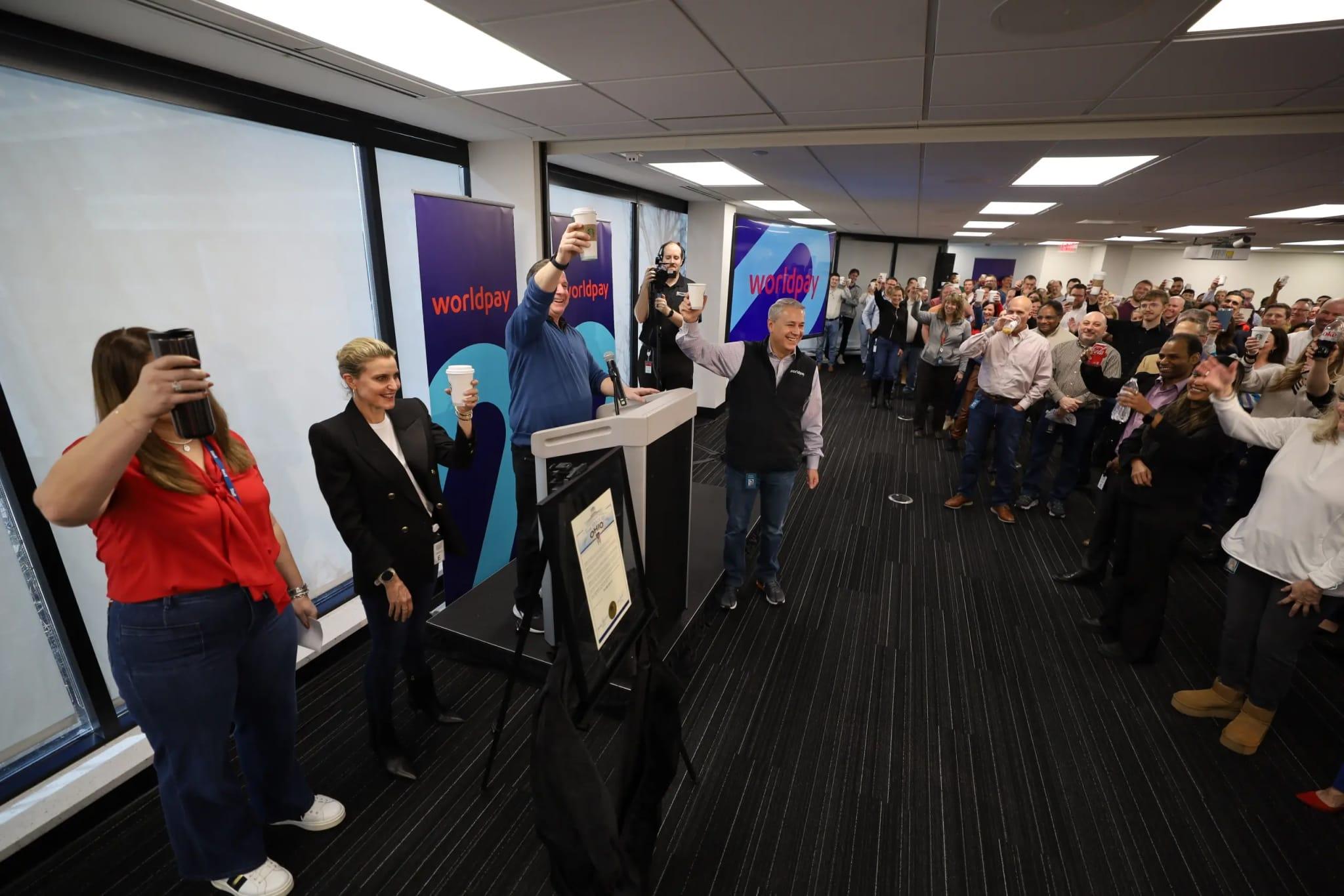 Wide angle of people in the office, holding up coffee cups in a cheers motion. Four people stand at a podium in front of everyone.