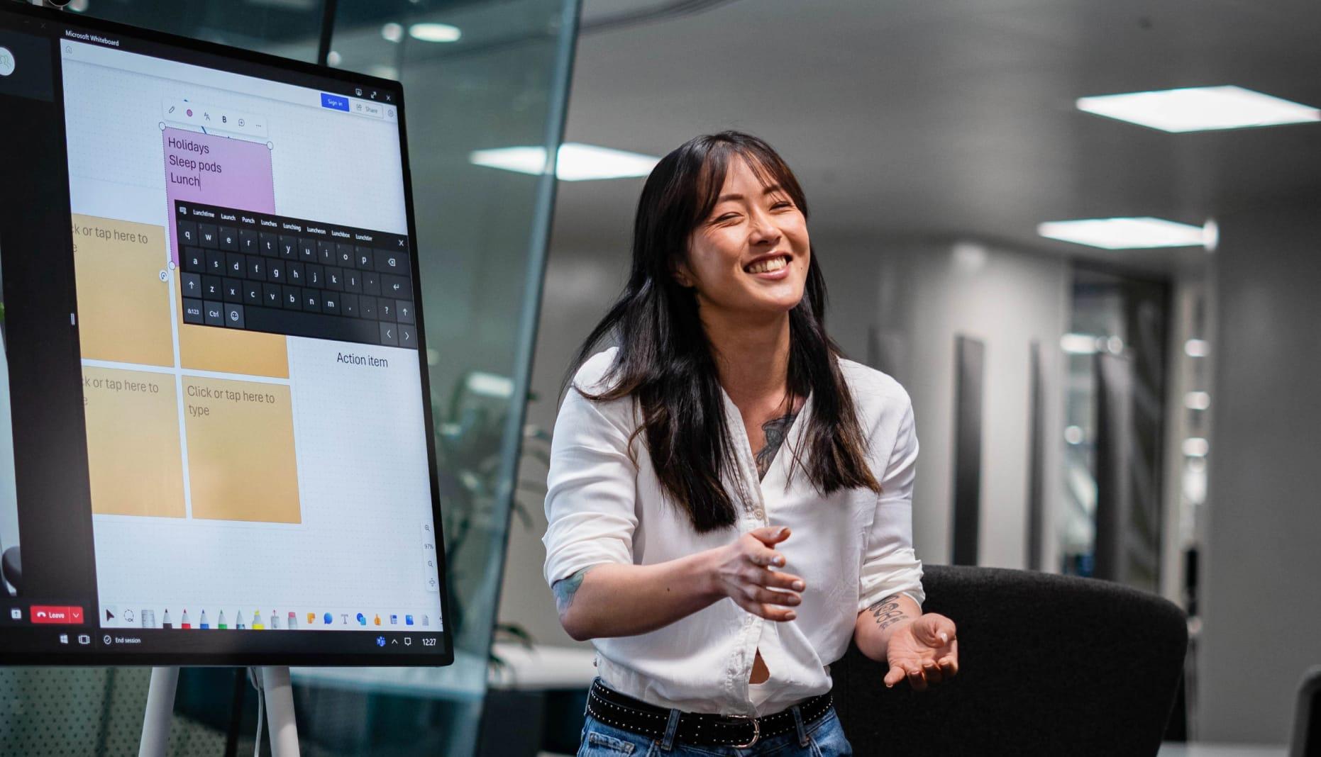 A woman is smiling whilst presenting in front of a screen in the office. She has long brown hair and visible tattoos and is wearing a white shirt.