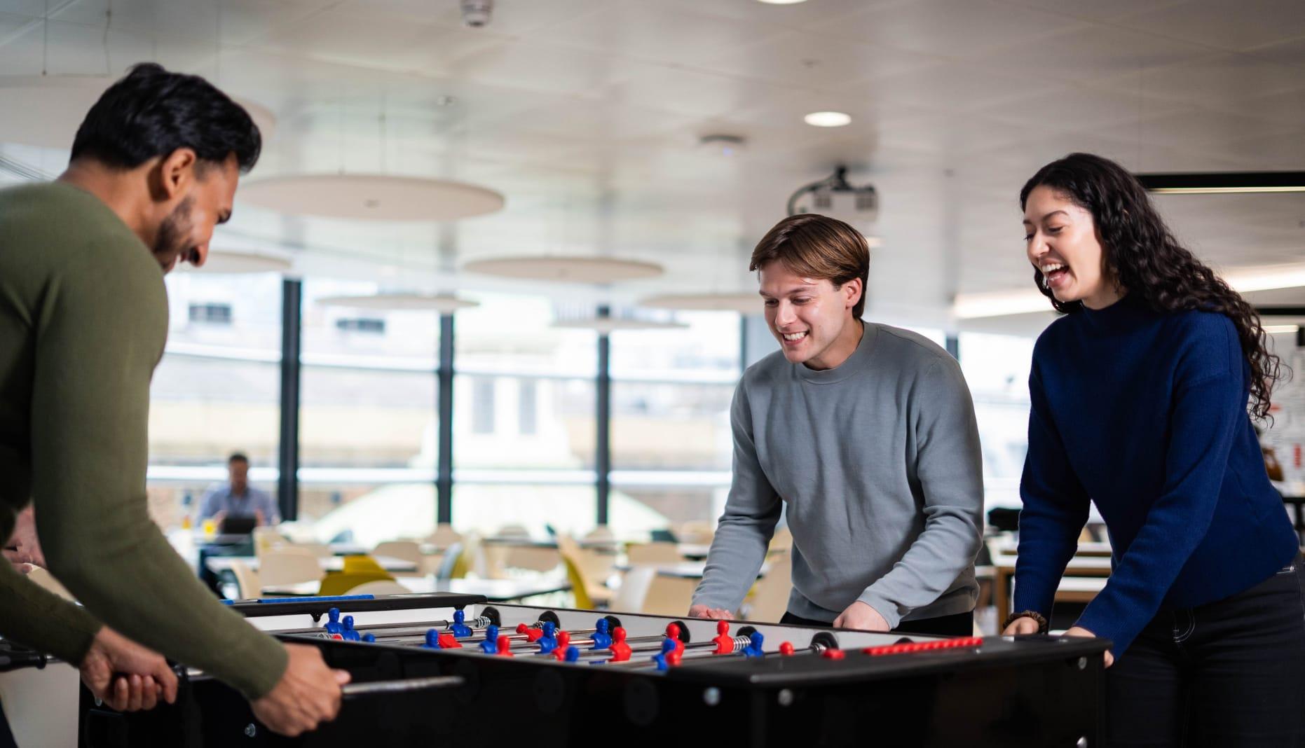 Three people play a game of table football in the office, in front of a large window.