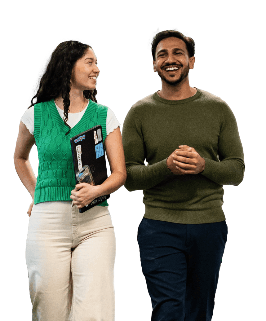 Two young people walking towards the camera in front of a blurred purple and photo background. The woman is wearing a green sweater vest, smiling and holding a book. The man is wearing a long-sleeved green top and laughing.