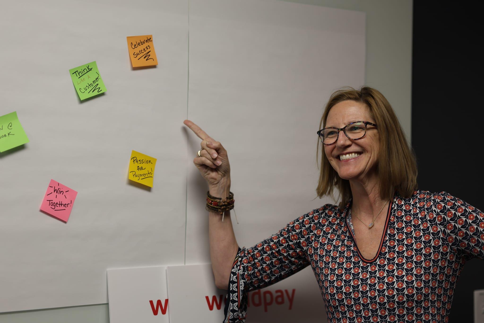 A woman in a patterned shirt smiles, pointing at a whiteboard that has yellow, green and pink sticky notes.