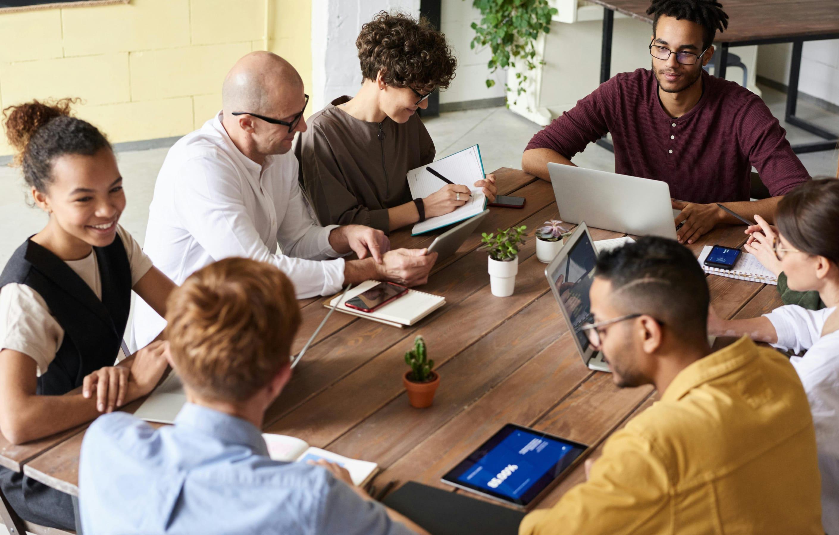 Five people are gathered around a wooden table with two small cacti. They are talking and taking notes with notepads and laptops.