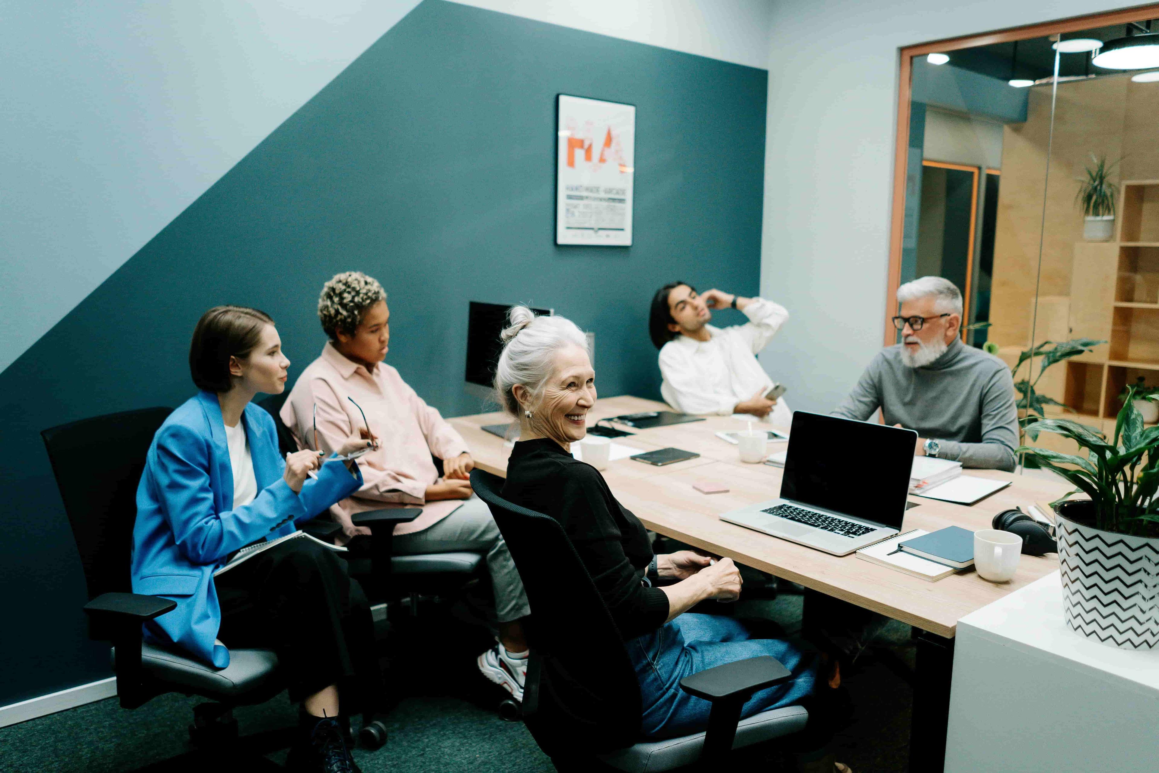 A group of people including a young and old demographic sat around a table in front of their laptops in an an office