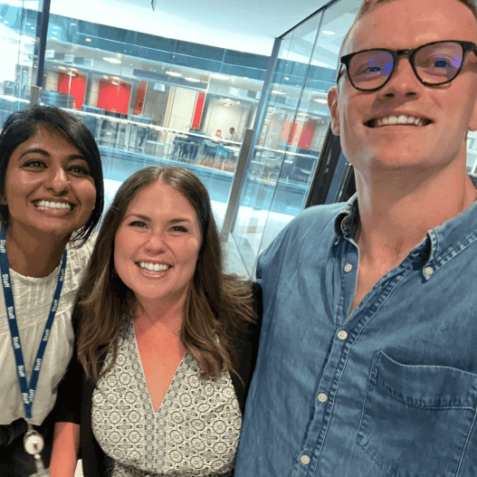 Three people smile for a selfie indoors, with glass walls and a busy office space in the background.
