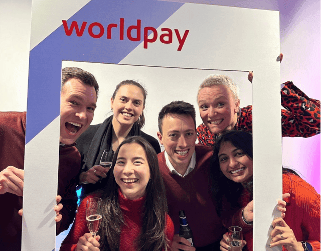 A group of people holding beer bottles and glasses of champagne pose with a cardboard picture frame that features the red Worldpay logo.