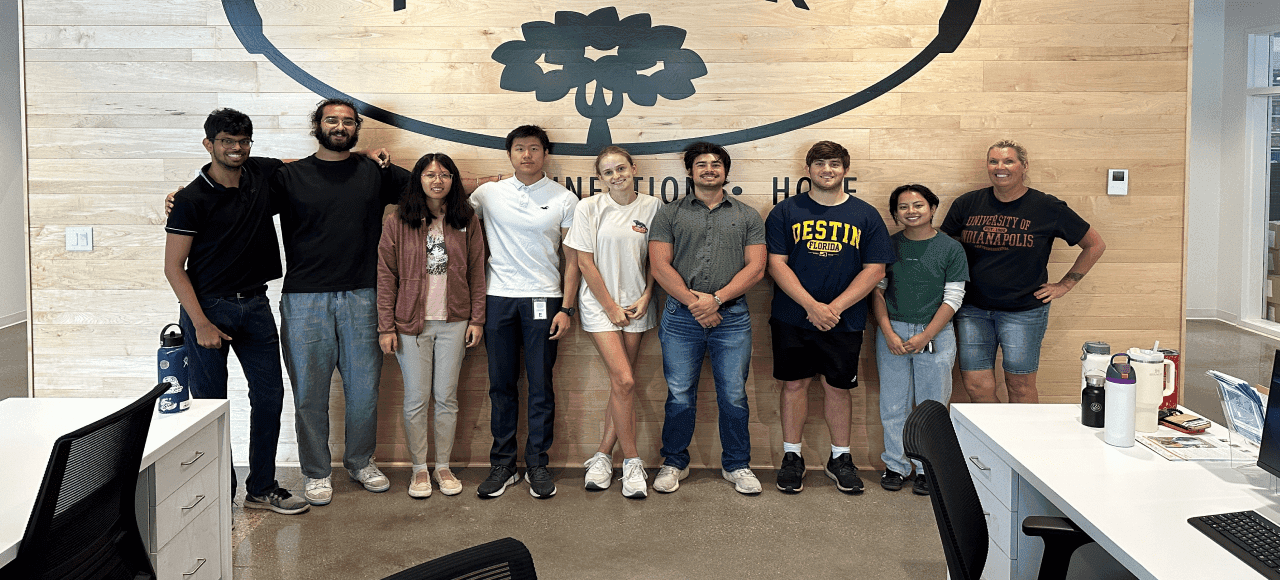 A line of nine people stand in front of an indoor wooden wall with a black logo on it.