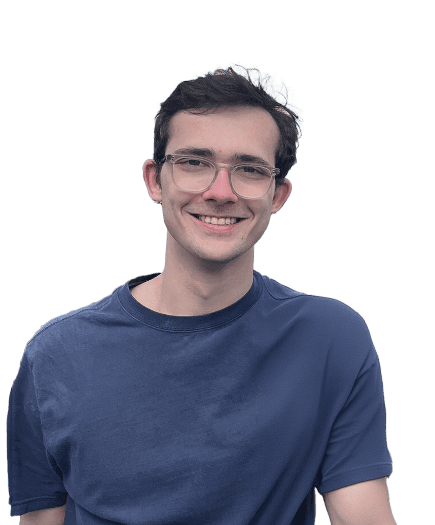 A man looks into the camera and smiles. He’s wearing glasses and a navy blue t-shirt and is in front of a blurred background.