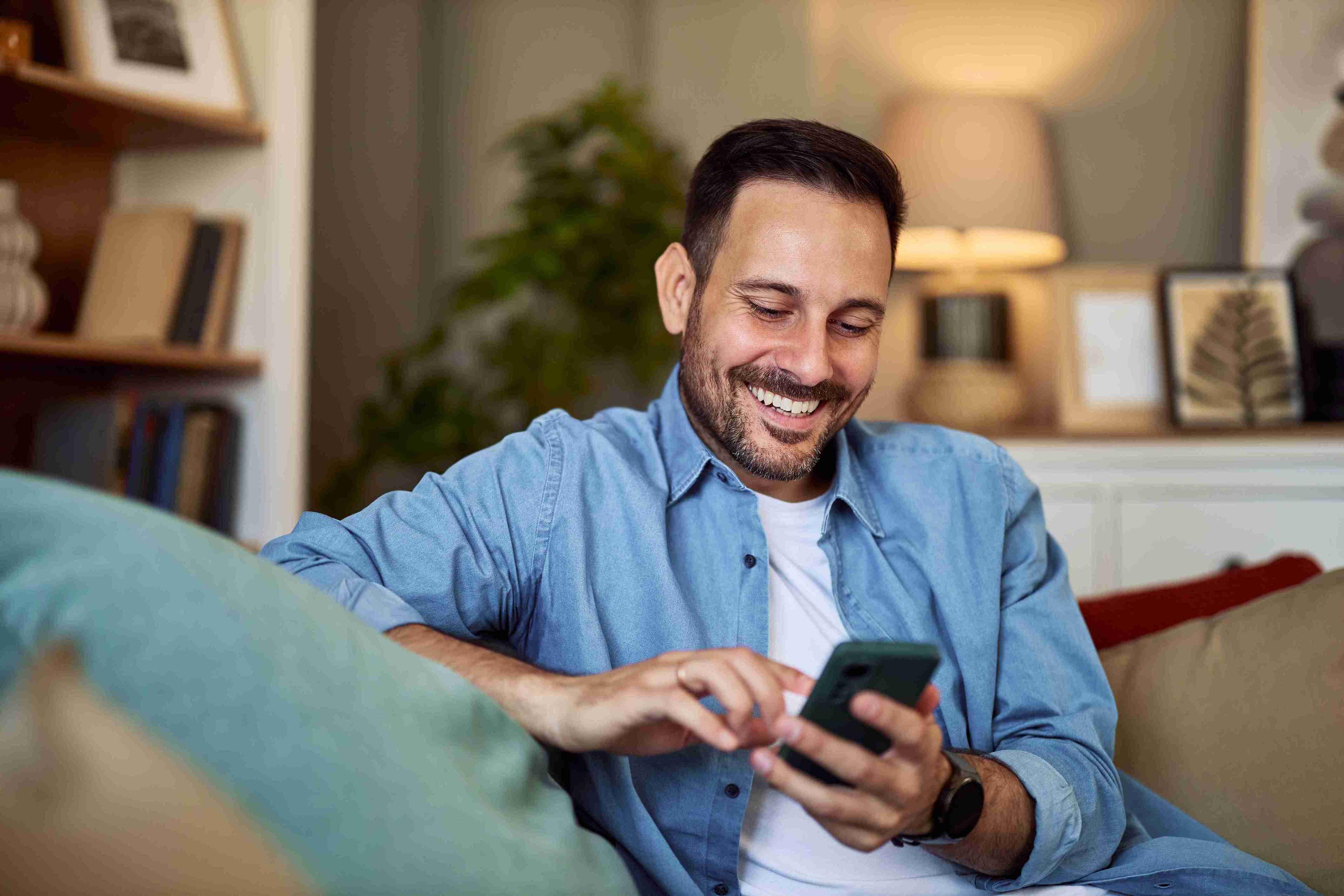 Man at home in blue shirt with white tshirt smiling looking at his phone