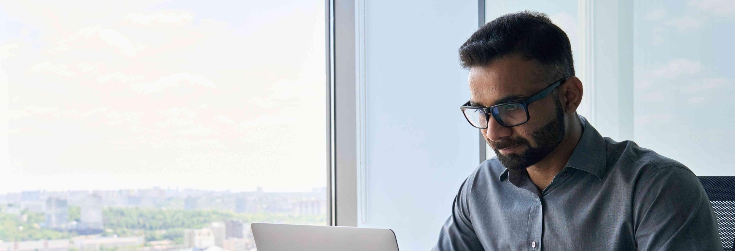 Man working on a silver laptop in front of a large window