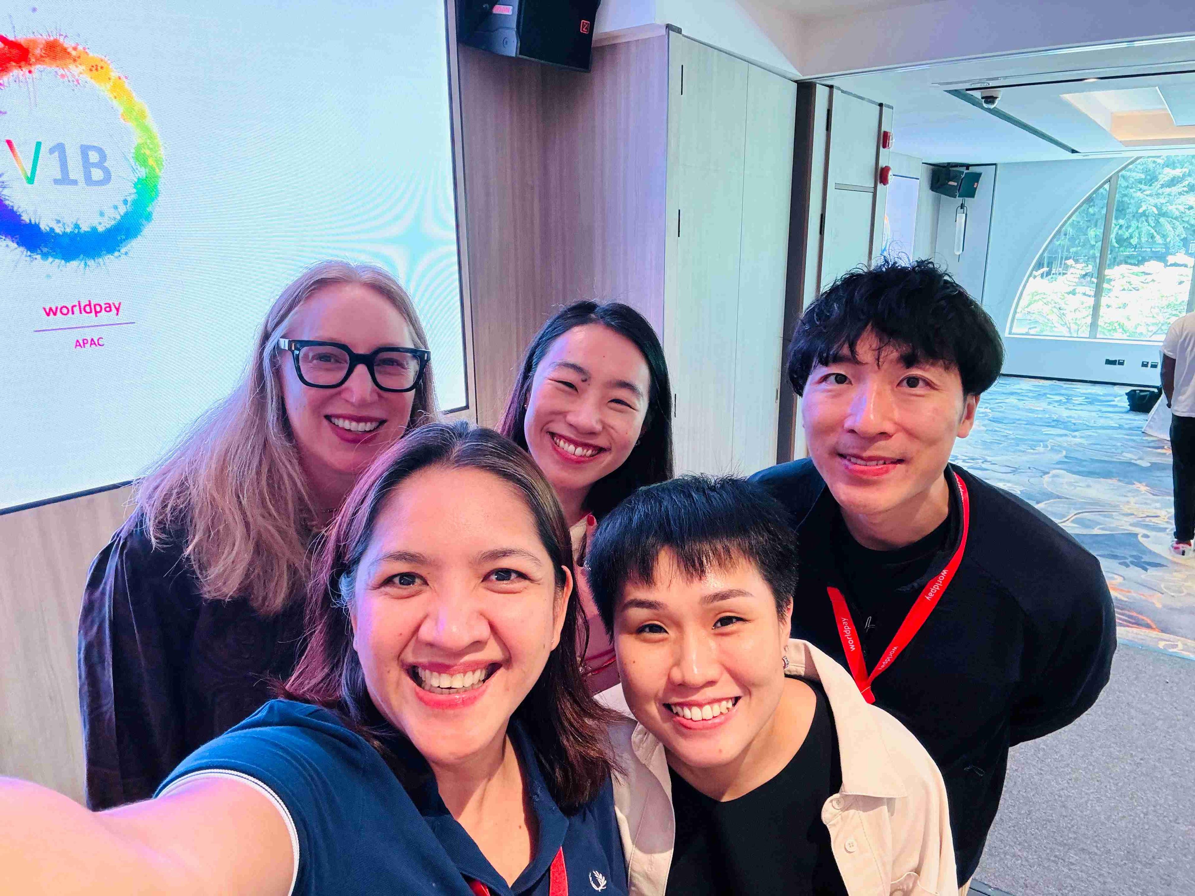 Four women and one man taking a selfie smile towards the camera in an office. A rainbow circle with ‘V1B’ in the middle is projected behind them on a whiteboard.