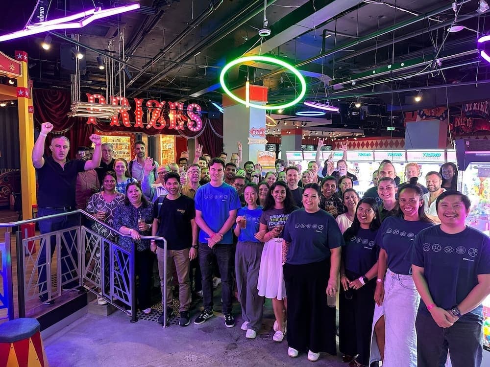 Large group of coworkers smiling in neon-lit arcade, some raising hands and a green ring light overhead and red sign behind.