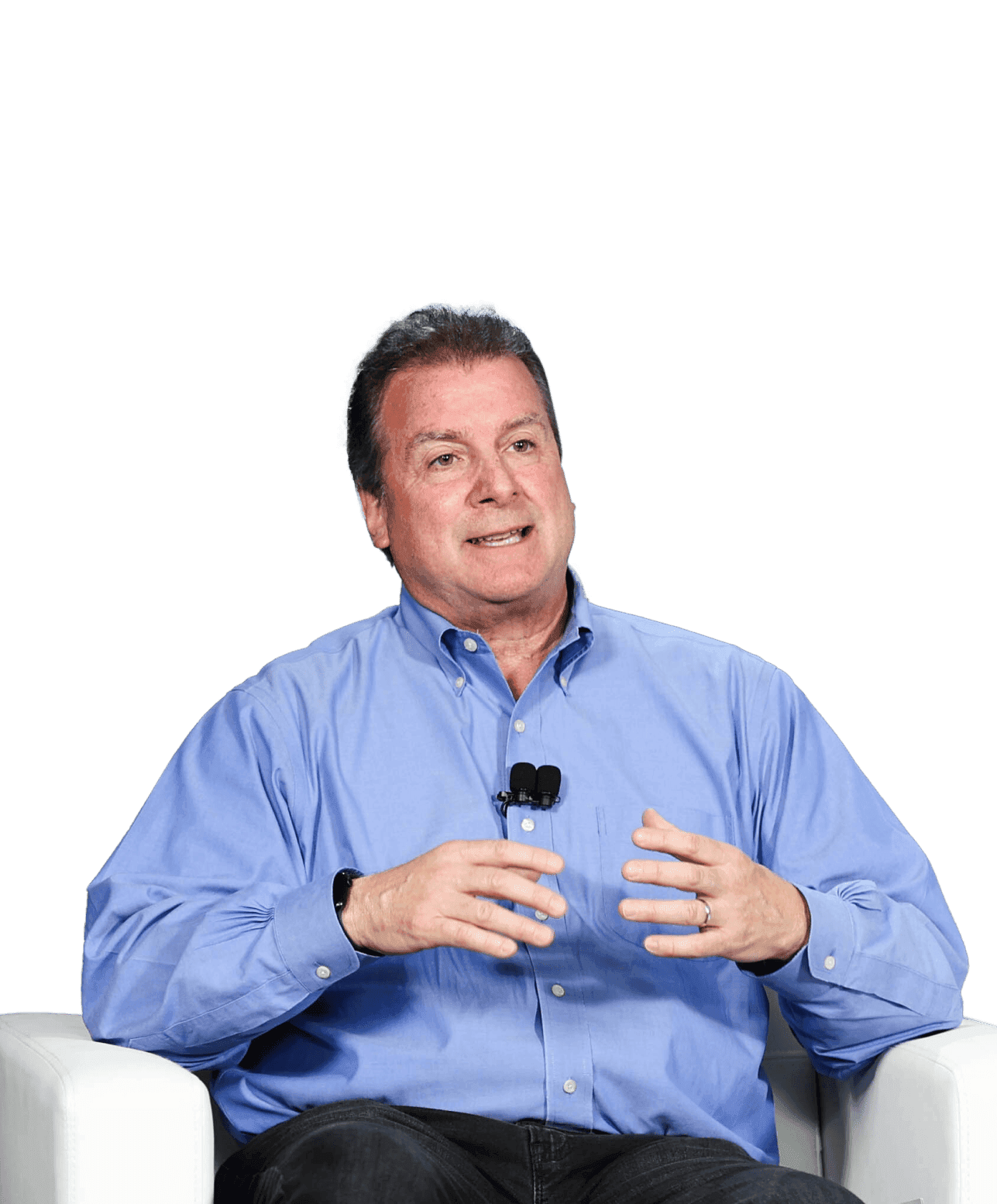 A brunette man in a blue shirt sits in a white chair, and the background is blurred. There’s a black lapel microphone attached to his shirt and he is mid-talking.