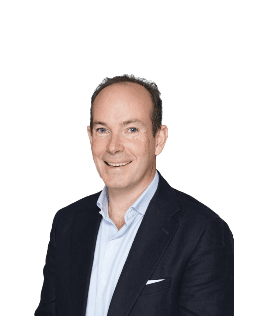 A brunette man in a white shirt and black blazer smiles into the camera, his office background is blurred.